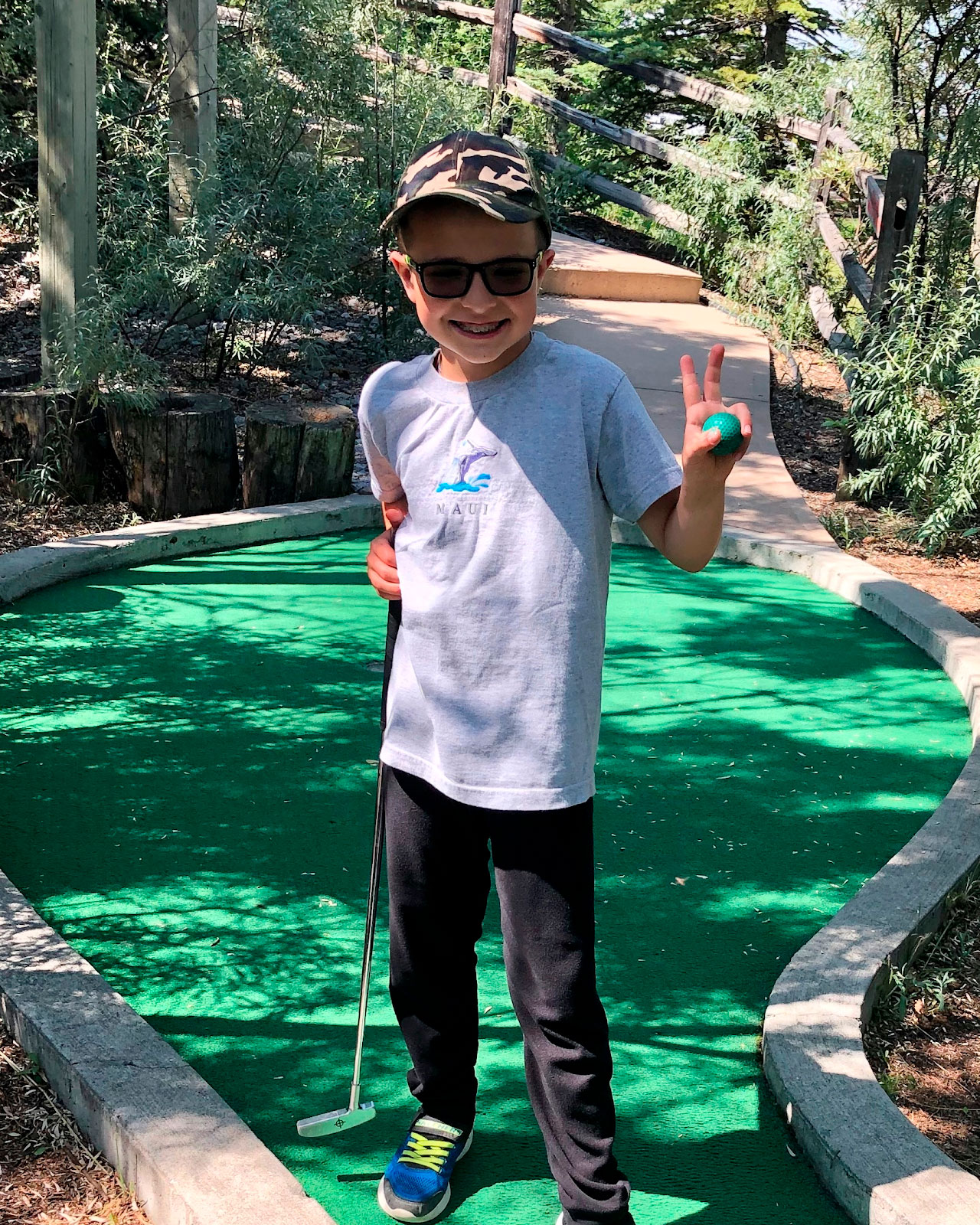 Jordan is standing on a mini golf course looking into the camera and smiling.  He is holding 2 fingers up as a peace sign with his right hand.