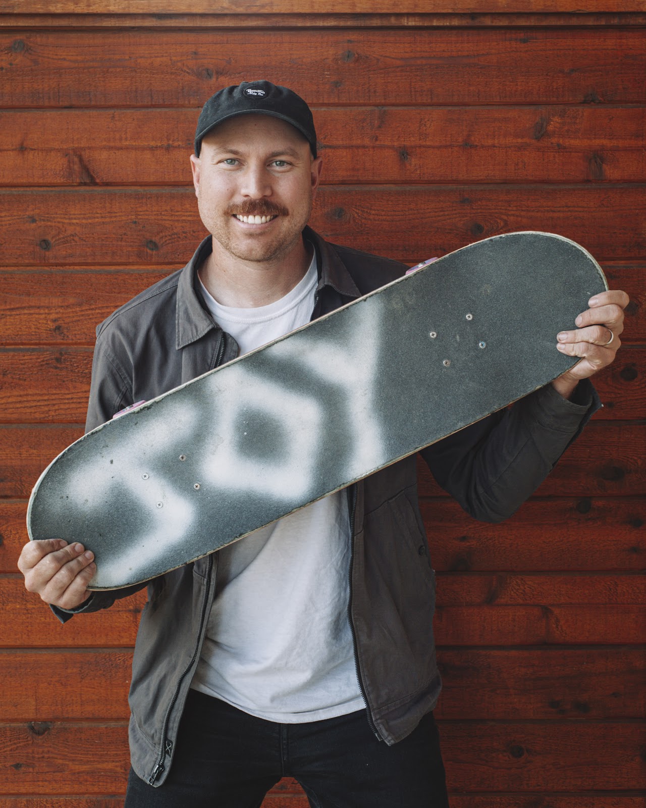 Matt standing smiling at the camera and holding his skateboard.  There is the word 